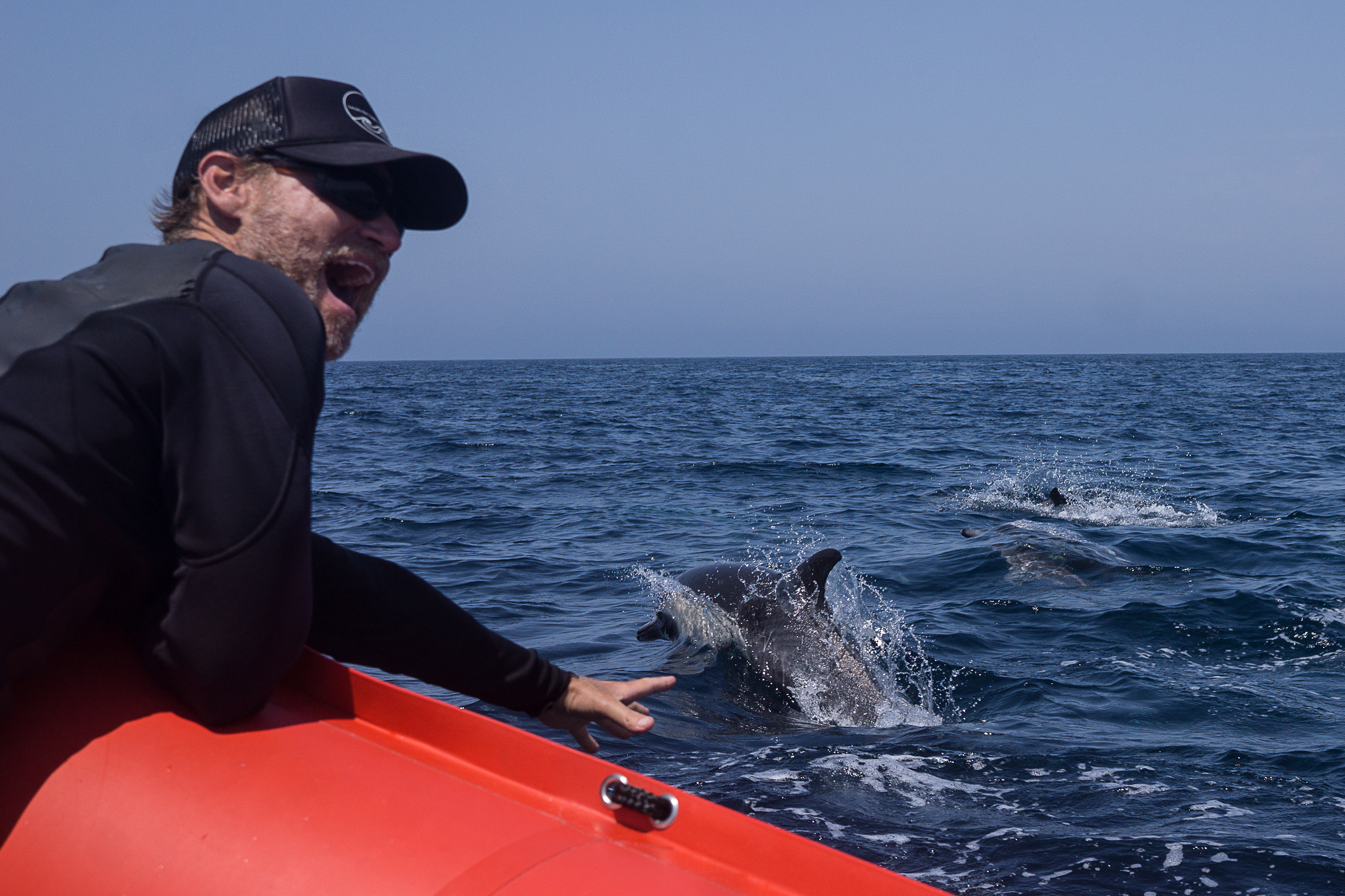 Rich German on red kayak reaching out toward dolphins splashing in dark blue ocean water