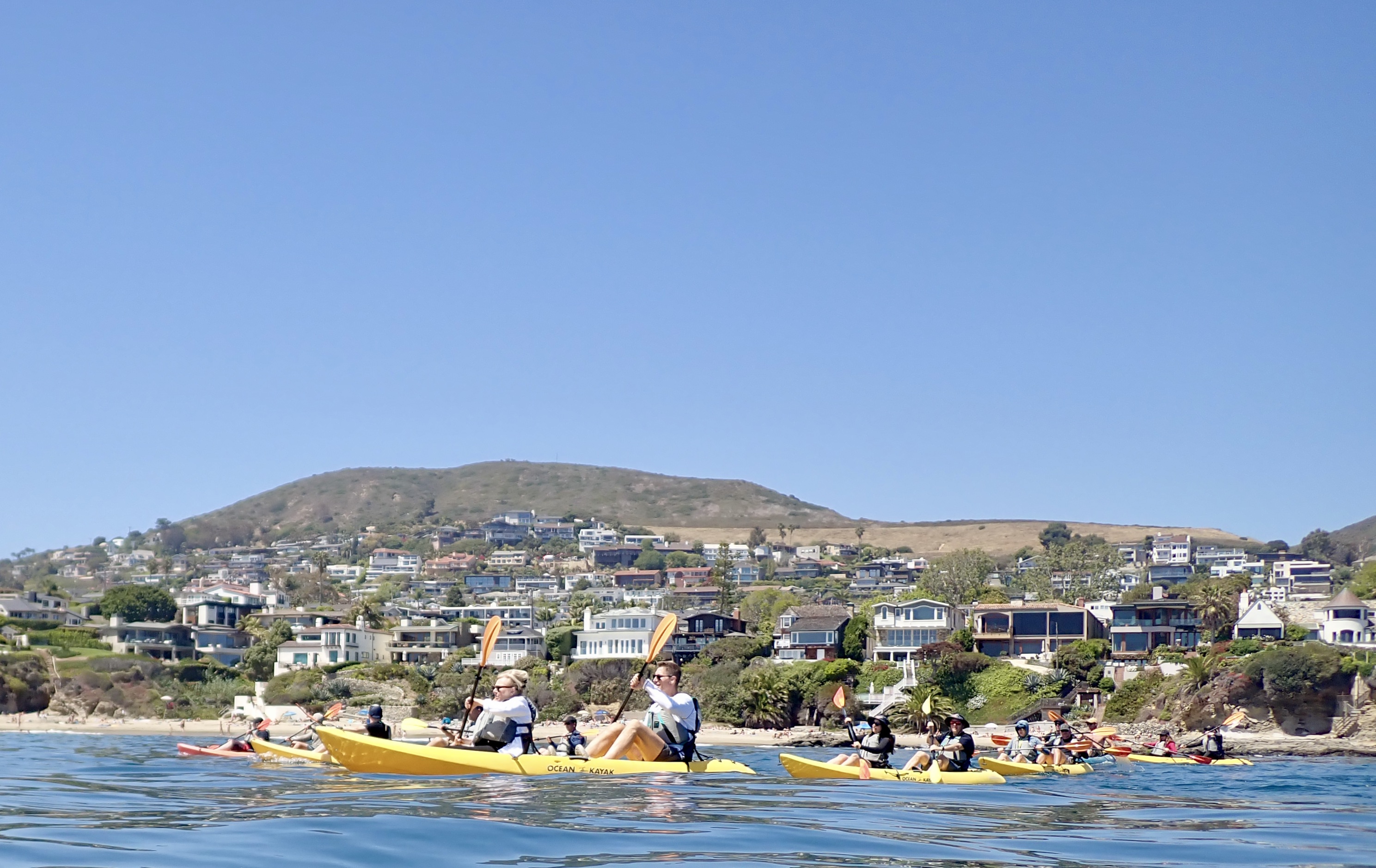 Kayakers paddling in calm blue waters with Laguna Beach coastline and hills in background